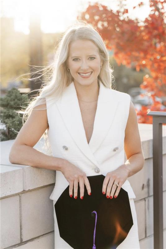 Blonde female smiling at the camera wearing a white dress and holding a law school regalia cap. She is leaning against a brick wall and the background is sunny with fall trees.