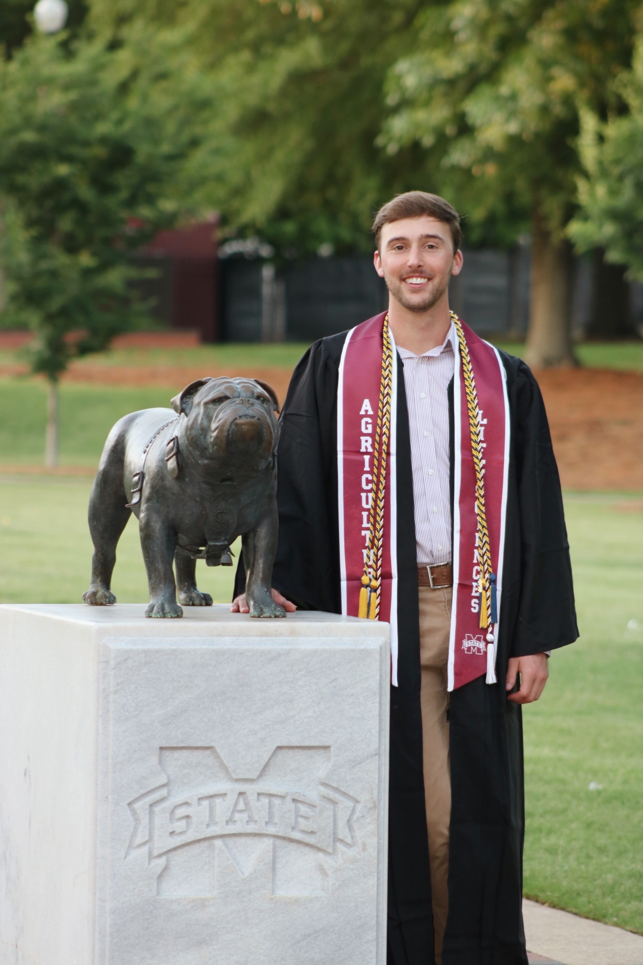  A man in a graduation gown stands proudly beside a bulldog statue, celebrating his academic achievement.
