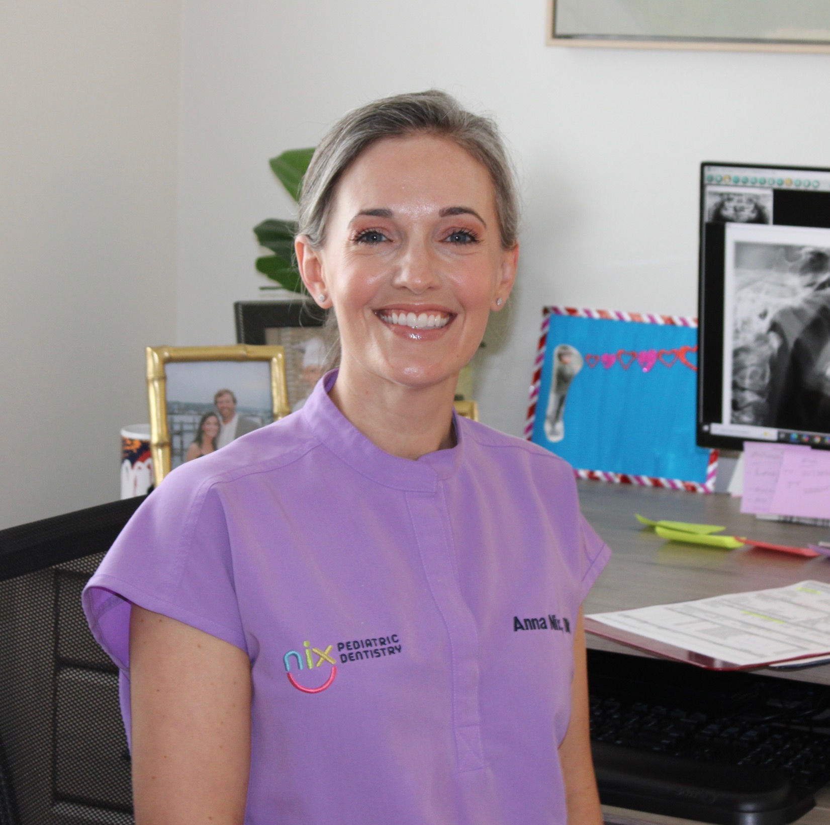 Smiling woman in a lavender dental uniform sits at a desk. She is in a dental office with framed photos and X-ray images on a monitor nearby.