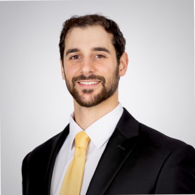 The image is a portrait image from the shoulders up of a man with dark hair and a bird wearing a suit with a yellow tie smiling at the camera. 