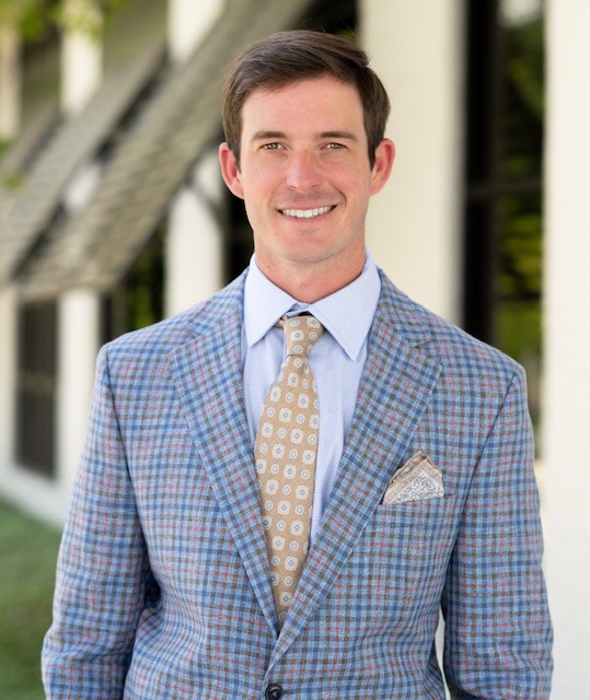 A man in a blue plaid suit, light blue shirt, and patterned tie stands outside in front of a building, smiling at the camera.