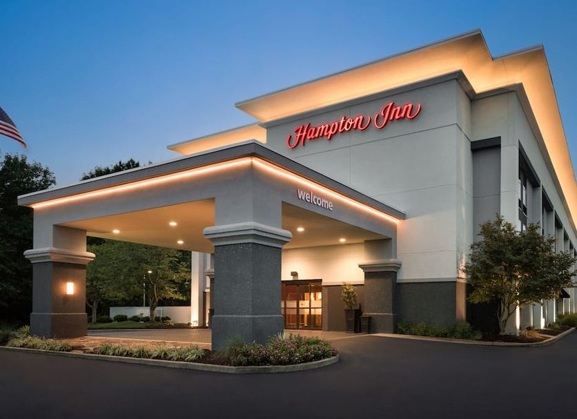 Hotel entrance at dusk with "Hampton Inn" sign in red above. Illuminated canopy and "Welcome" signage create a warm, inviting atmosphere.