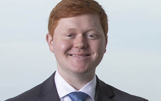 Man with red hair smiling at the camera wearing a dark grey suit, white top, and blue patterned tie. The background is light blue. 