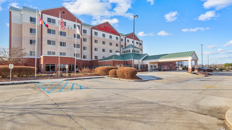 Five-story hotel with red brick and beige exterior, green roof, and entrance canopy. Flags wave out front under a sunny sky with fluffy clouds.