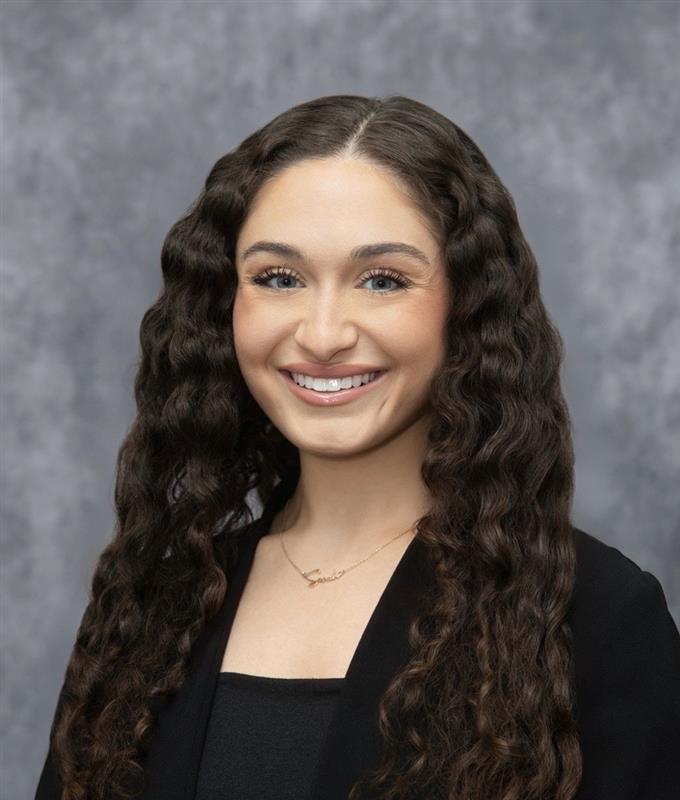 Headshot style image of woman with curly dark hair smiling at the camera. She is wearing a black jacket and a black shirt. The background of the image is grey. 
