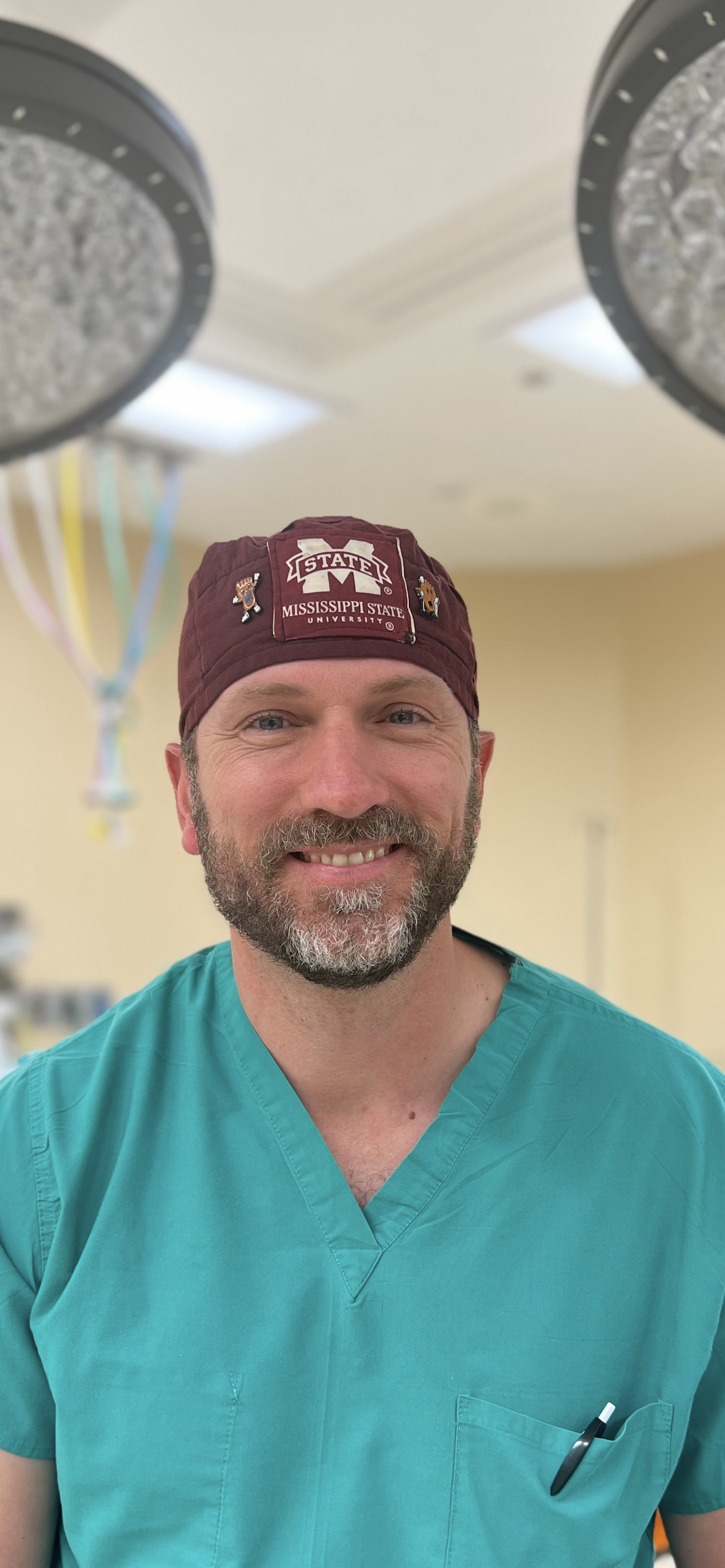 Image of man from waist up smiling at the camera. He is wearing blue scrubs and a maroon scrub cap that has a Mississippi State logo and says "Mississippi State University". The background is a blurred surgery room. 