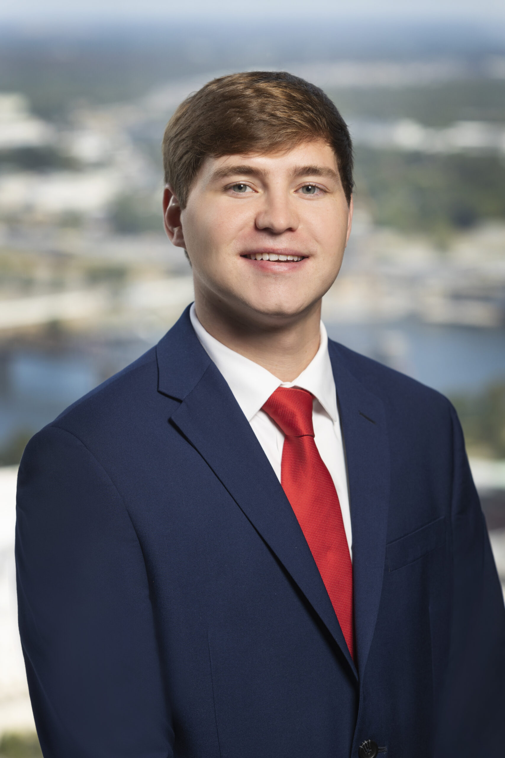 Man with brown hair smiling at the camera. He is wearing a navy suit, white top, and red tie. The background is a blurred image of a river and buildings. 