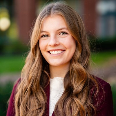 a woman with curly brown hair smiling at the camera. She is wearing a maroon blazer and white top with a blurred background of a brick building and grass. 