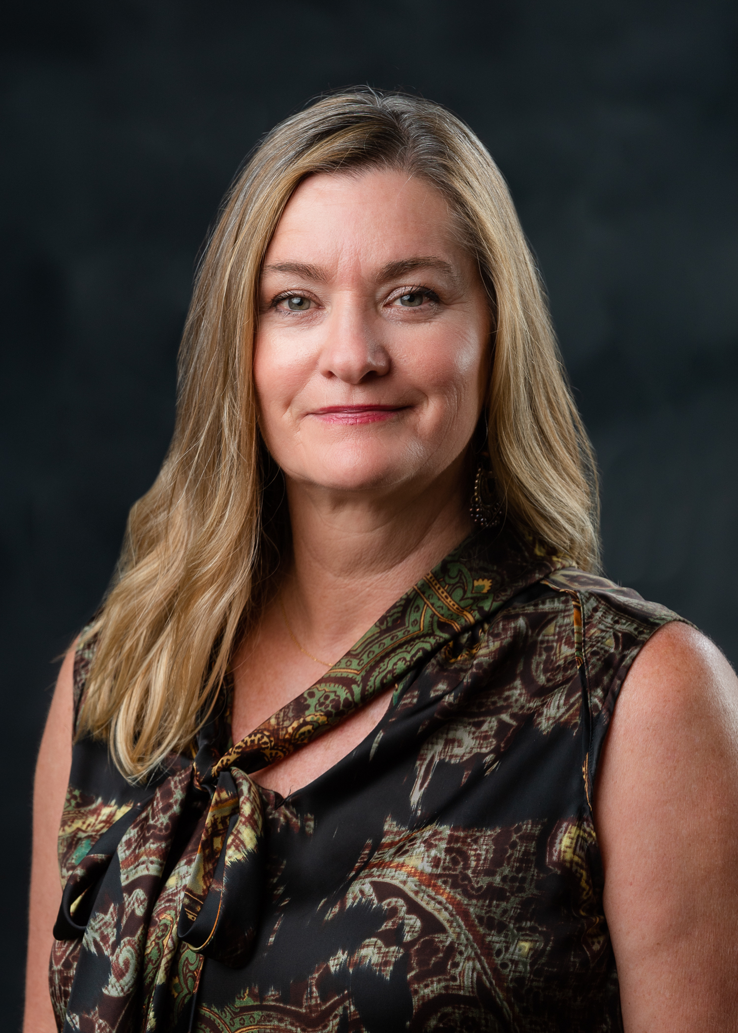 headshot style image of woman with blonde hair looking at the camera. She is wearing a green, maroon, and black patterned top. the background is a dark grey. 