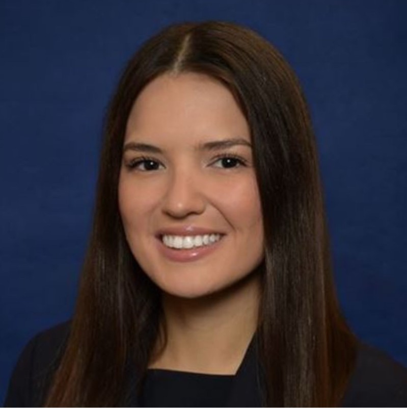 Woman with long dark brown hair smiling at the camera. She is wearing a black blazer and top. The background is navy blue. 