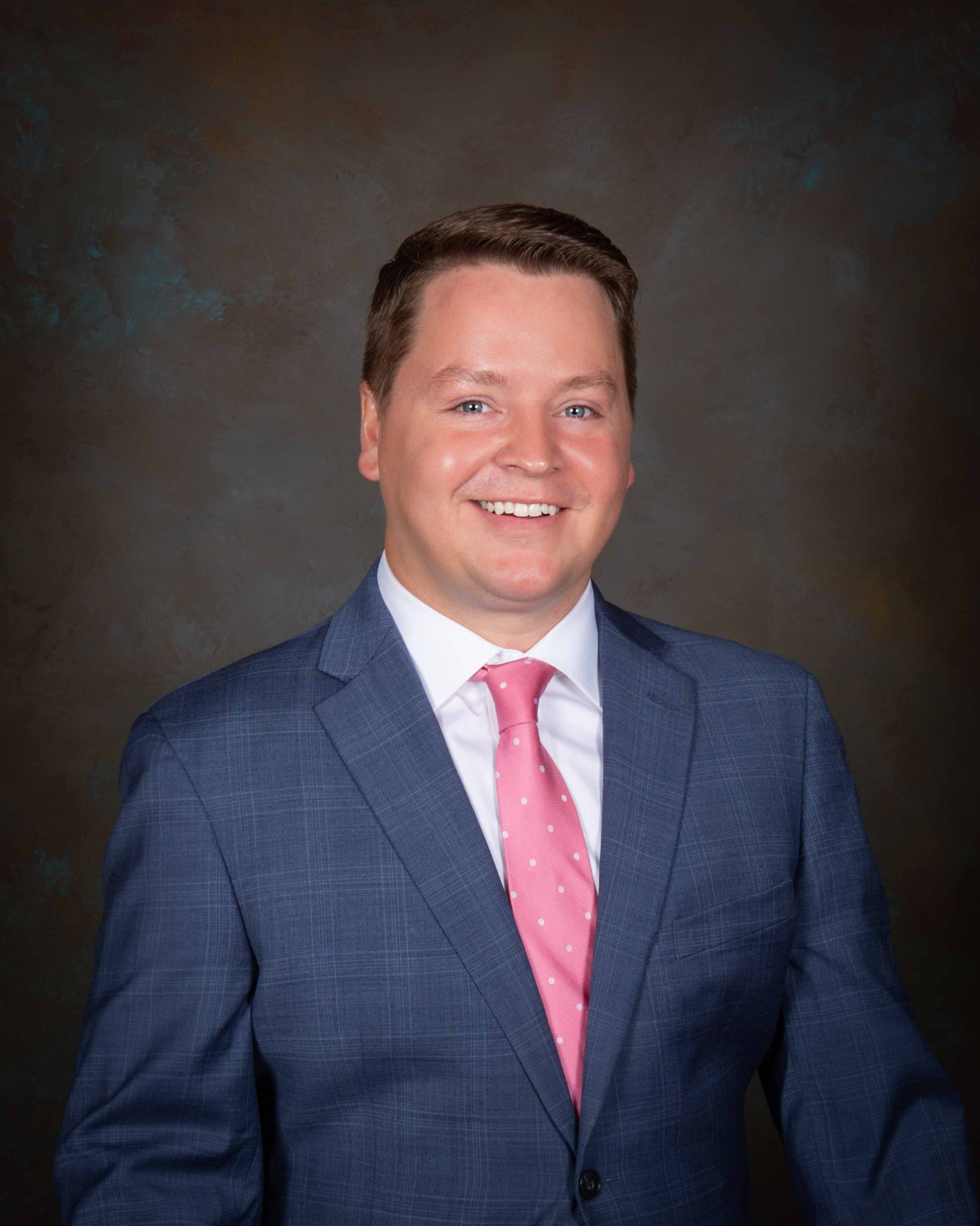 Smiling individual in a blue checkered suit and pink tie against a dark, textured background. Professional and approachable demeanor.