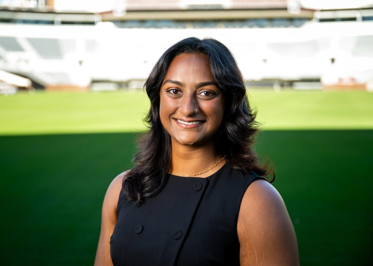 Smiling woman in a black dress stands in front of a sunlit, empty sports stadium. The mood is confident and professional with a bright backdrop.