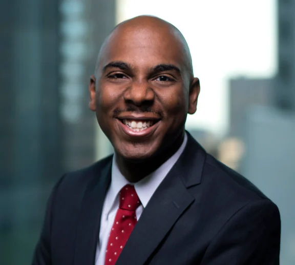 A man wearing a navy suit, blue shirt, and red and white polka dot tie smiling at the camera with a blurred background. 