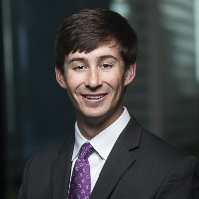A man with dark hair wearing a dark grey suit, white shirt, and purple and blue patterned tie smiling at the camera with a blurred background. 