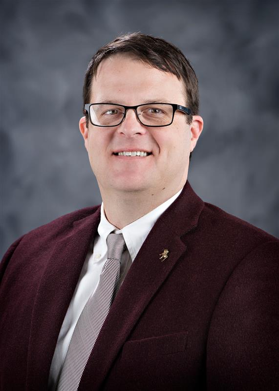 Headshot style image of man with dark hair and glasses smiling at the camera. He is wearing a maroon suit jacket, white shirt. and a maroon striped tie. The background is dark grey. 