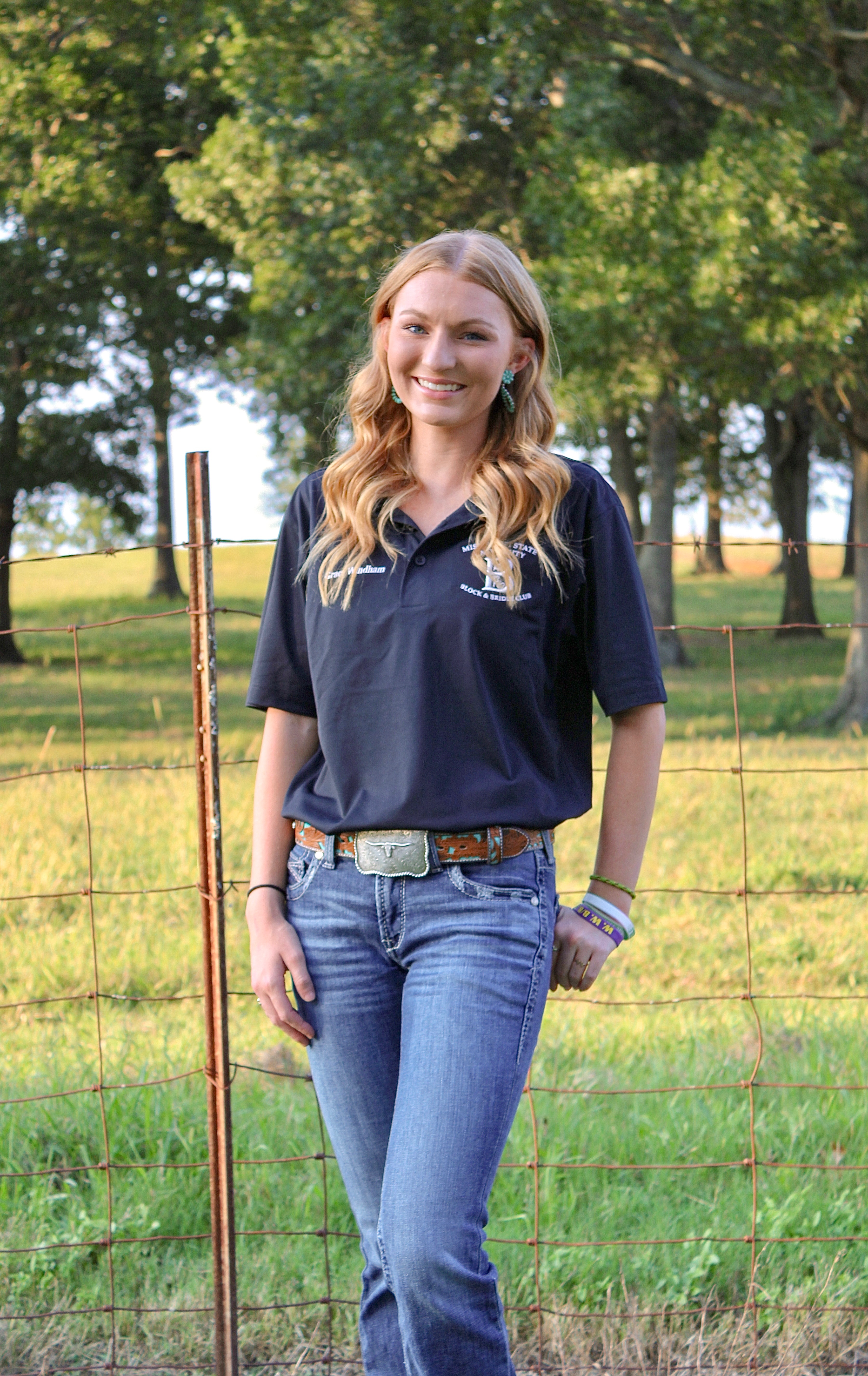 Photo is a full image of a female with long blonde hair smiling at the camera. She is wearing blue jeans, a belt, and a black polo that reads "Grace Windham" on the left and "Mississippi State University Block and Bridle on the right. Behind her is a fence, grass, and trees. 