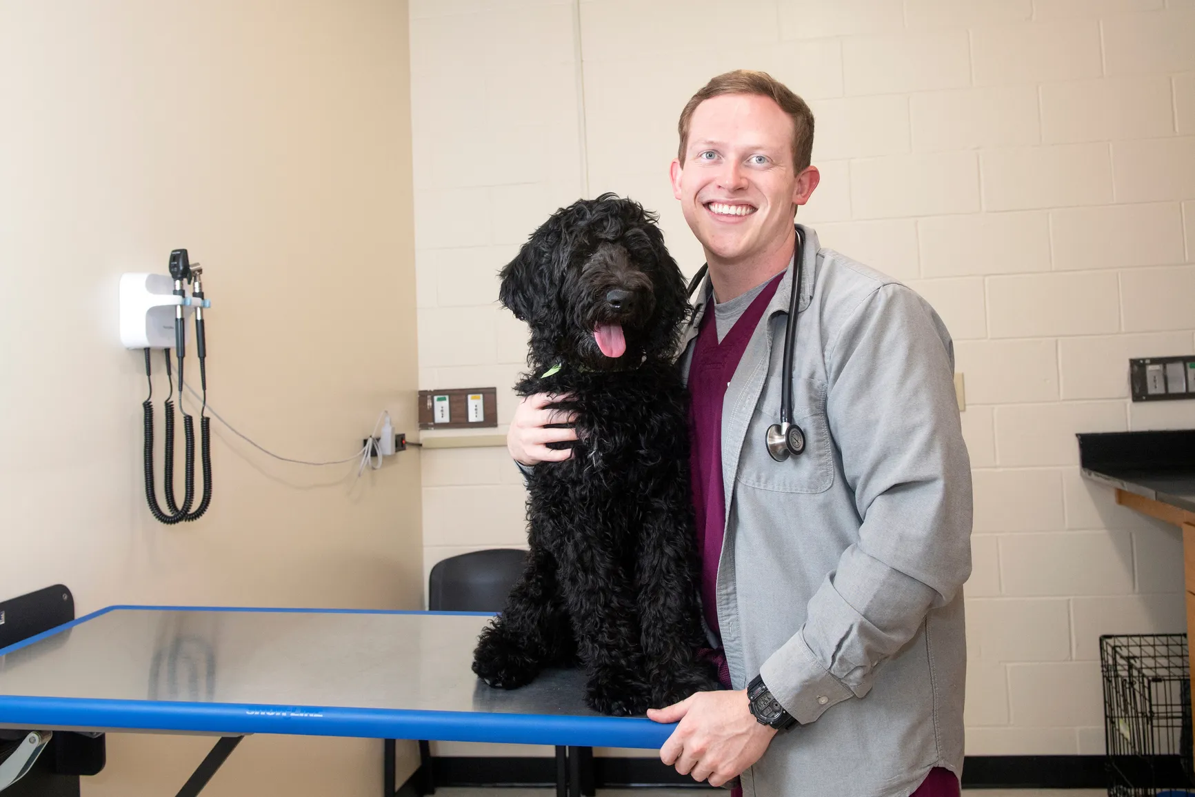 Image of a man wearing a grey scrub jacket and maroon scrubs smiling at the camera. He has a stethescope around his neck and has his right arm wrapped around a black dog with curly hair that is sitting on an examination table. The background is a vet office with medical equipment. 