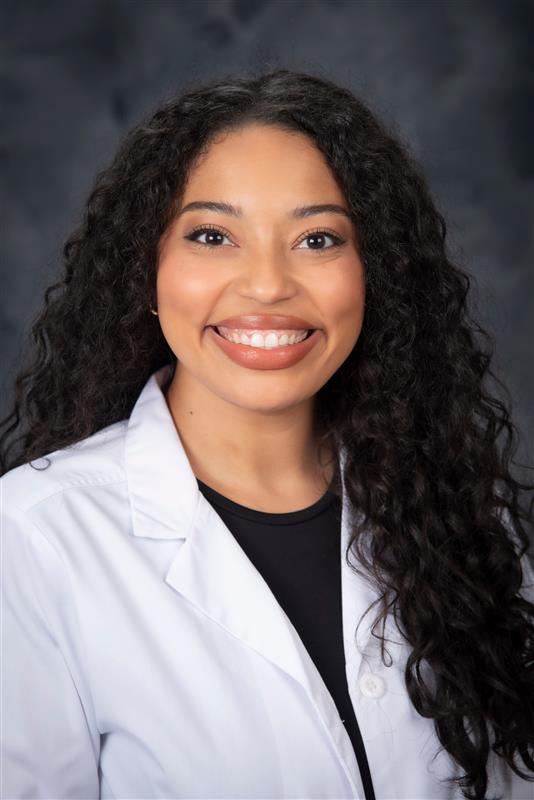 Photo of a woman smiling at the camera with long, dark, curly hair wearing a white lab coat with a dark shirt underneath. The background is gray and it is a portrait style shirt. 