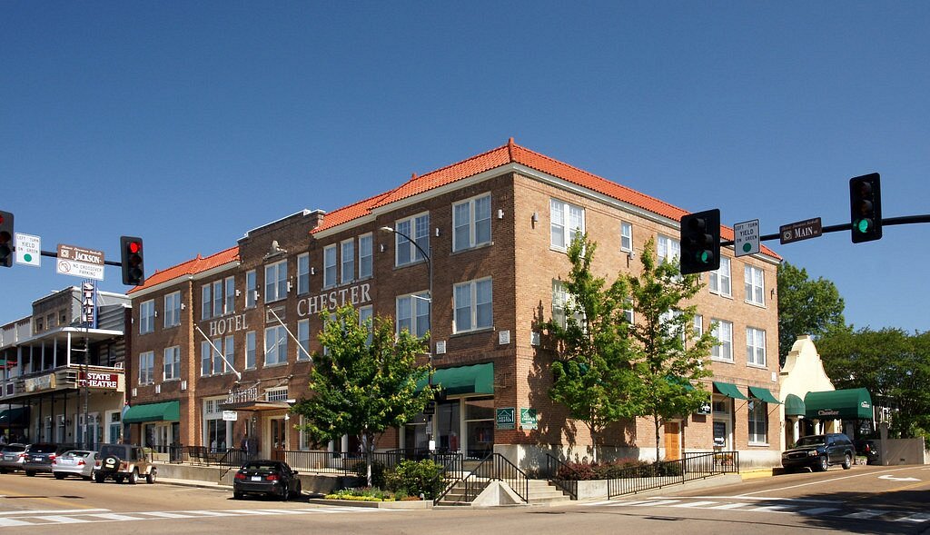 Historic three-story Hotel Chester, with red-tiled roof and green awnings, on a sunny corner. Traffic lights and cars complete the classic scene.