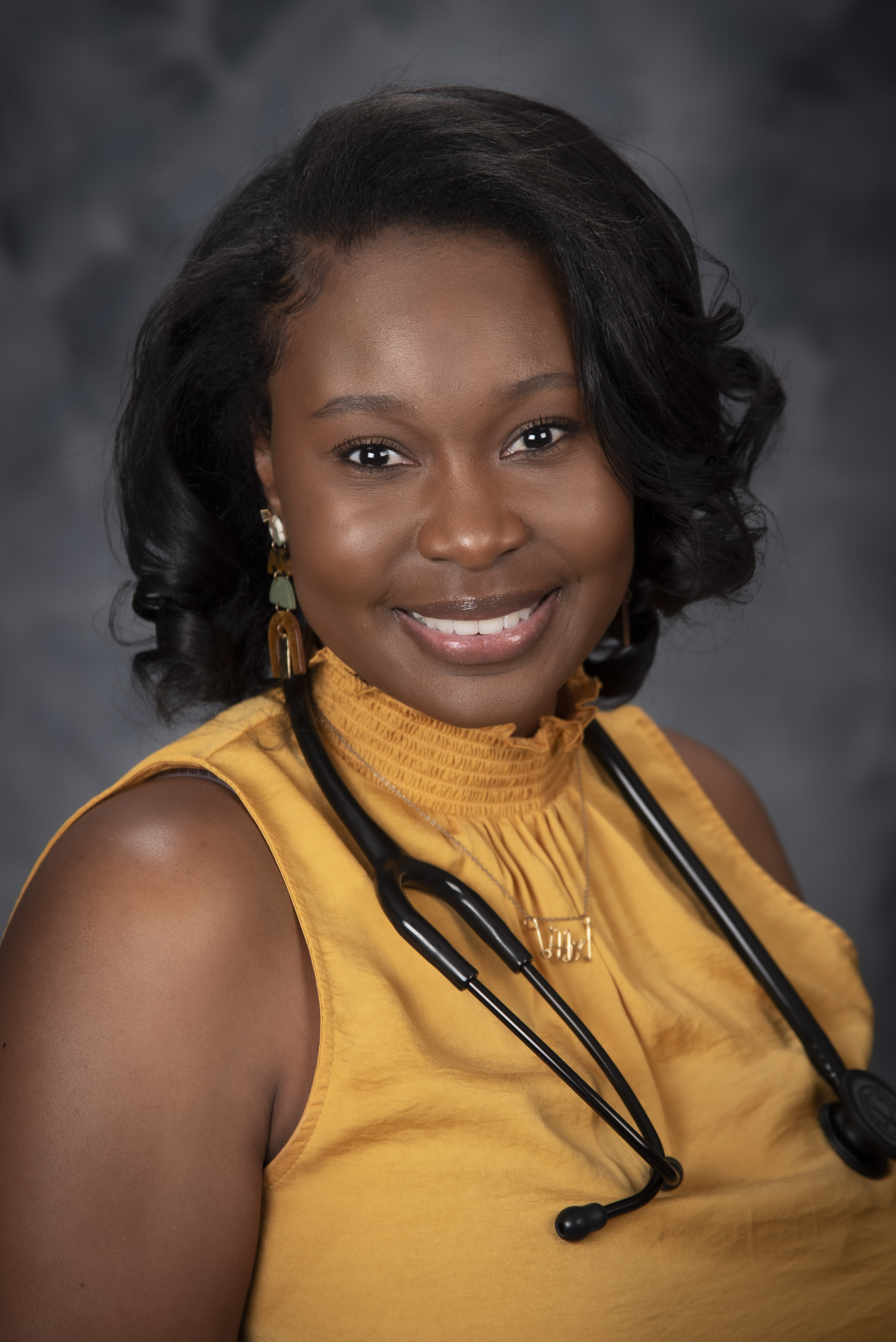portrait style image of woman wearing yellow dress top with a stethoscope around her neck smiling at the camera. the background is a plain grey backdrop. 