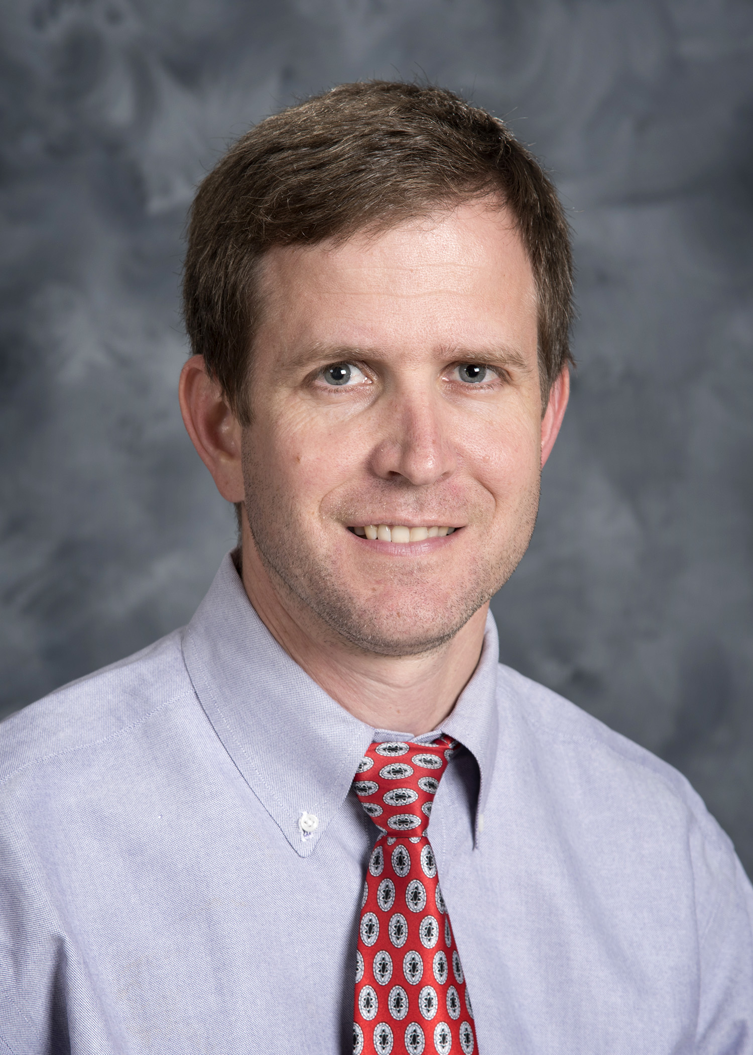 Headshot style image of man with brown hair smiling at the camera. He is wearing a blue button down shirt and a red tie that has white and blue ovals on it. The background is dark grey. 