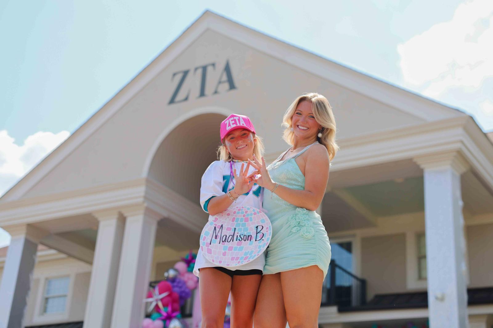 Two smiling women stand in front of a building with "ZTA" on it. One wears a pink cap