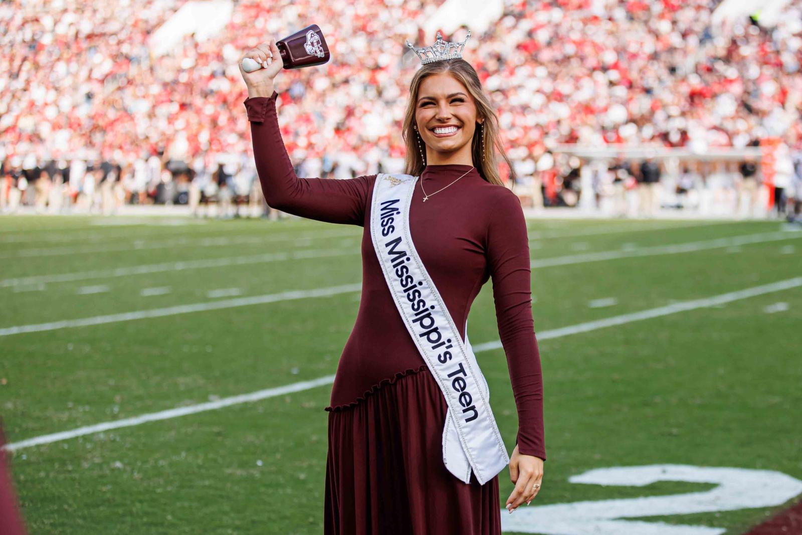 A smiling woman wearing a crown and a "Miss Mississippi's Teen" sash stands on a football field
