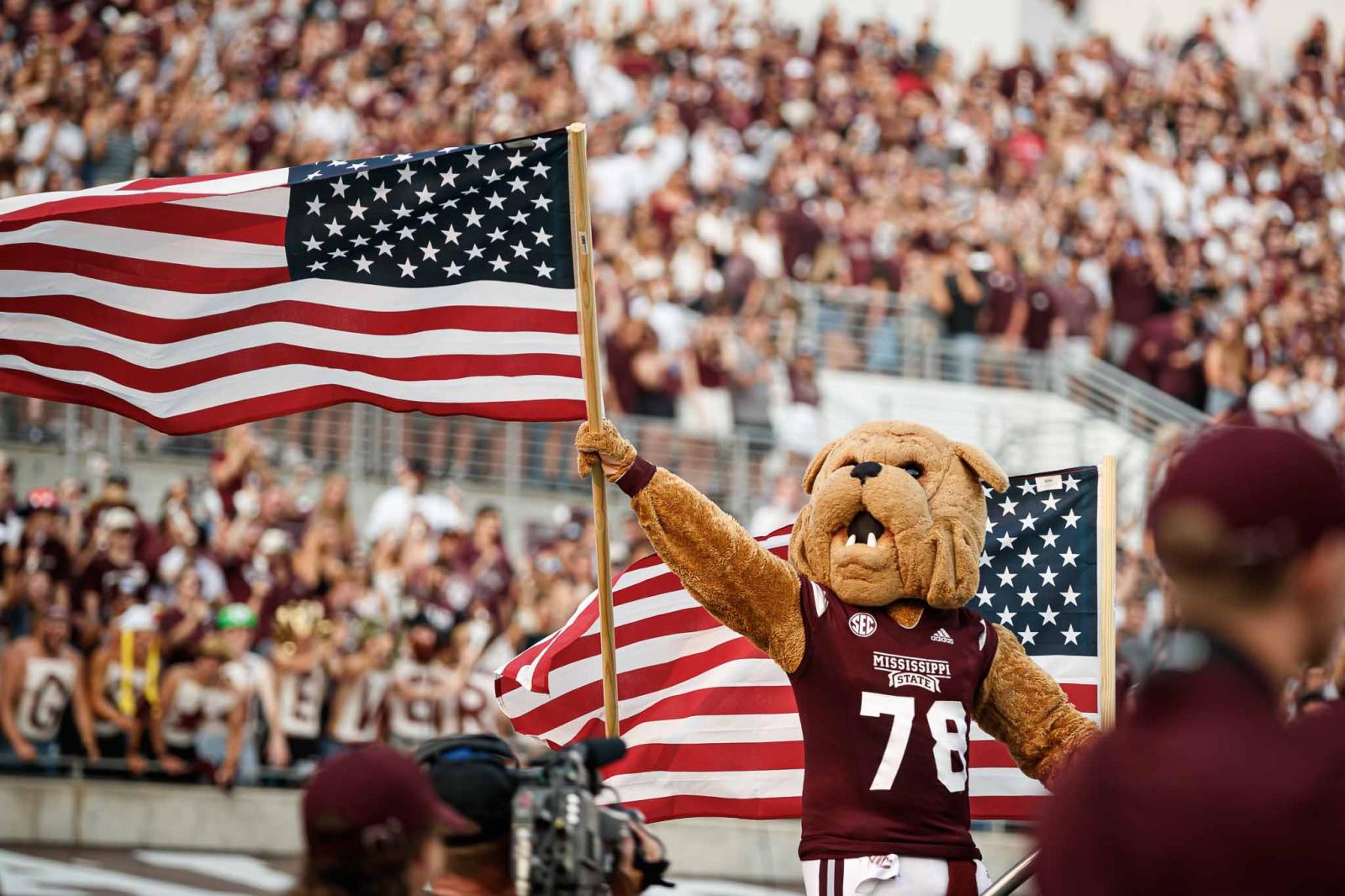 Mississippi State University Bully mascot holding an American flag in front of a full Davis Wade stadium.