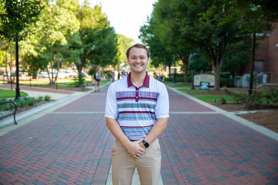 Eli Myrick, Admissions Counselor, standing on the brick road between Montgomery Hall and McCool Hall smiling in a maroon and white shirt